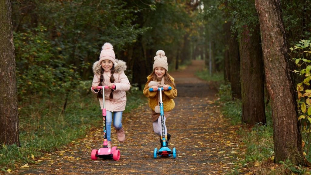 Two girls wearing beanies and winter jackets ride scooters on a leaf-strewn path in an autumn park.